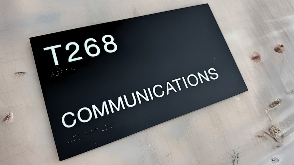 Black ADA-compliant room identification sign with raised white tactile lettering reading "T268 Communications" for interior office wayfinding.