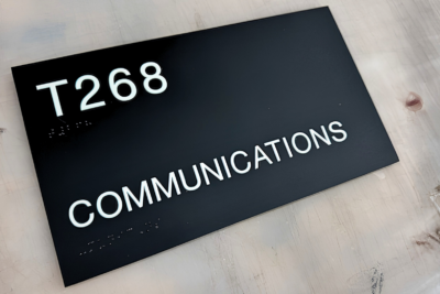 Black ADA-compliant room identification sign with raised white tactile lettering reading "T268 Communications" for interior office wayfinding.
